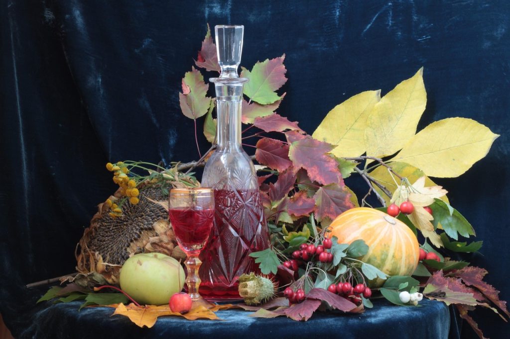 A still life arrangement featuring a crystal decanter and glass filled with red liquid showcases the art of wine decanting, surrounded by autumn leaves, a sunflower head, an apple, yellow squash, berries, and a dark blue draped background.
