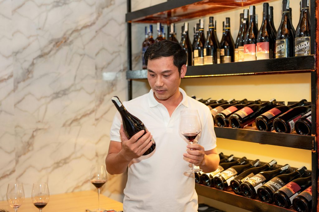Asian man in a wine cellar holding a wine bottle and glass, considering his choice.