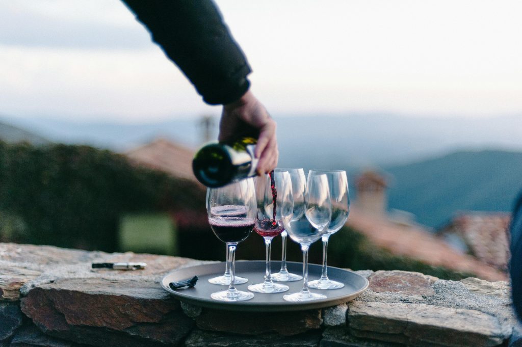 Hand pouring red wine into glasses on a tray in an outdoor setting with hills in background.