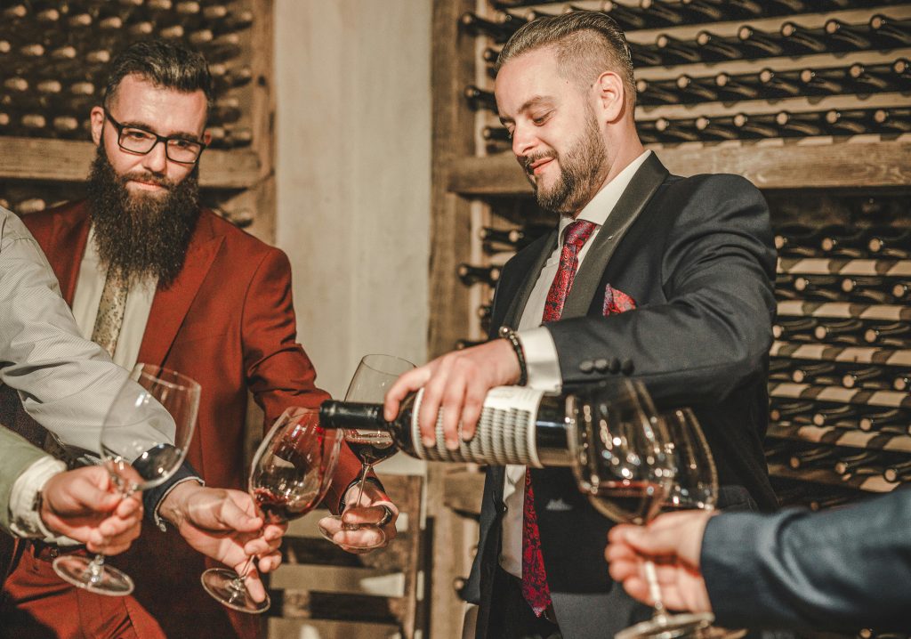 Two men in suits share wine at a stylish cellar gathering.