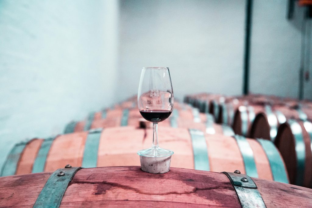 A glass of red Madeira wine sits elegantly on an oak barrel in a Stellenbosch winery cellar.