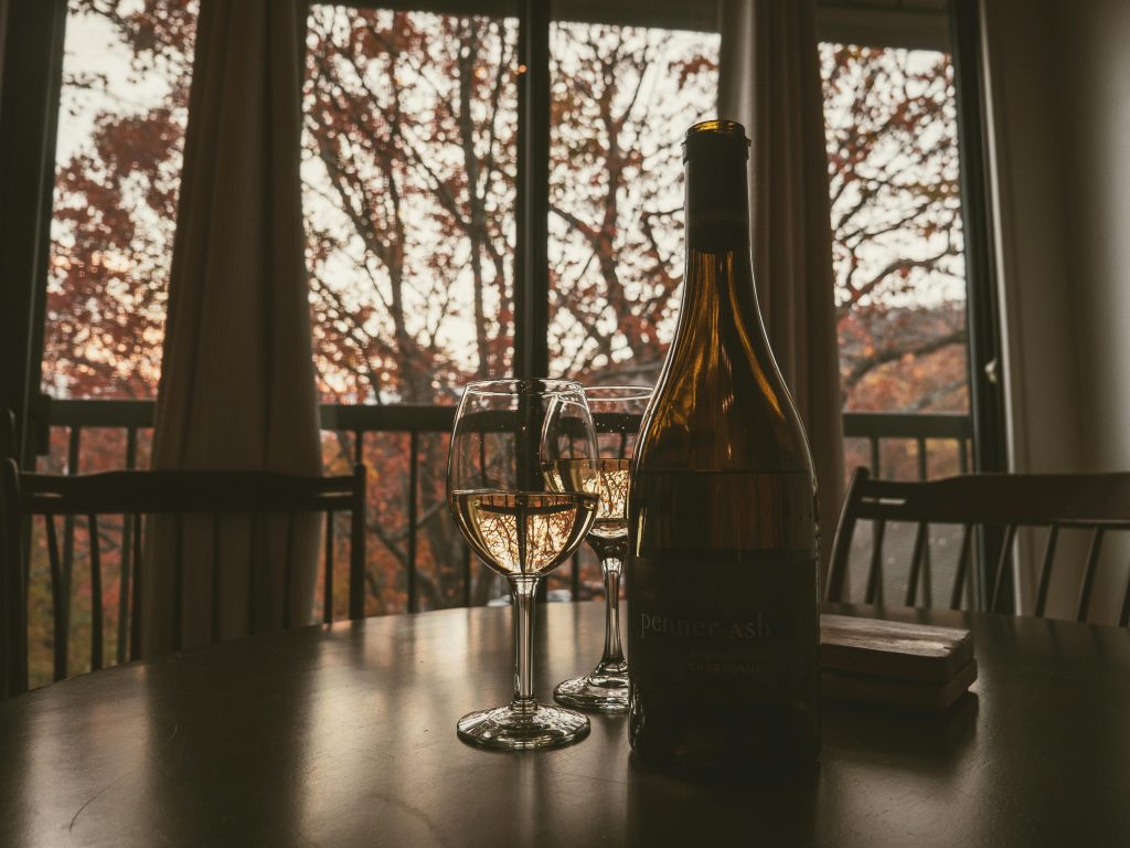 How to Prepare Your Wine Fridge for Seasonal Changes A warm, cozy setting with two glasses of white wine and a bottle indoors, framed by autumn trees.