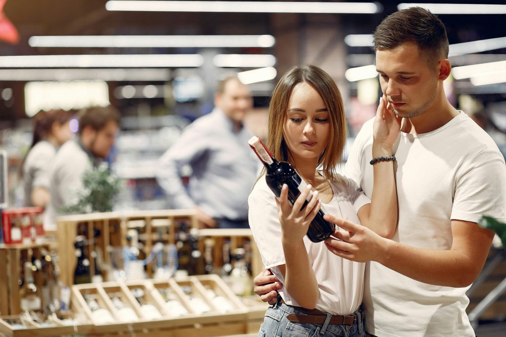 Young man and woman in casual outfit hugging and looking on bottle of wine while choosing alcohol for lunch in contemporary store together