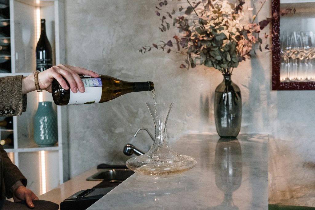 A hand pouring white wine into a decanter on a stylish bar counter with a decorative vase.
