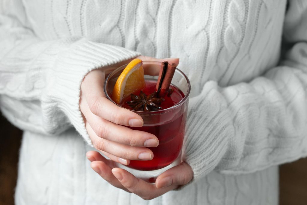 Warm mulled wine with spices and citrus, held by hands in a cozy sweater, perfect for winter relaxation.