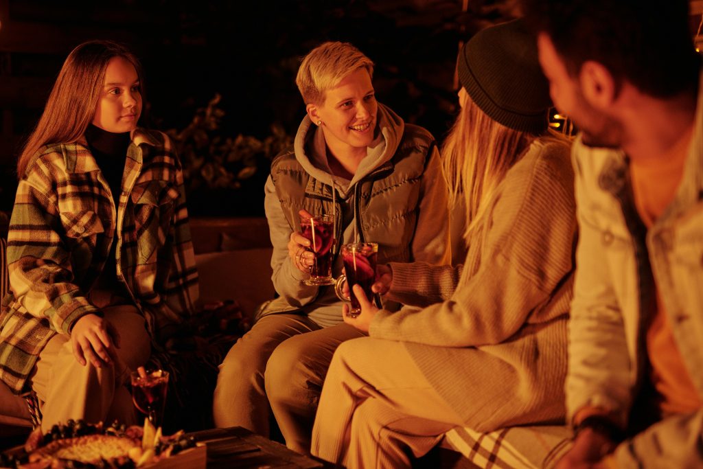 Group of friends gathering around bonfire in night garden having warming drinks and chatting in leisure