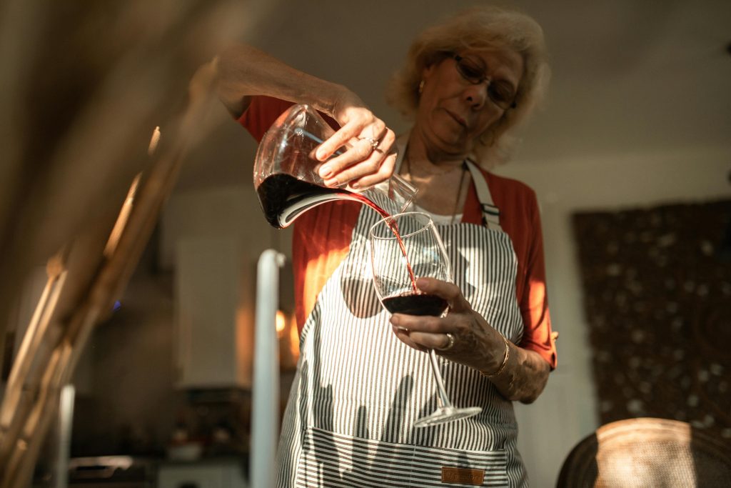 An older woman wearing glasses and a striped apron pours red wine from a decanter into a glass in a warmly lit kitchen, elegantly demonstrating the art of wine decanting.