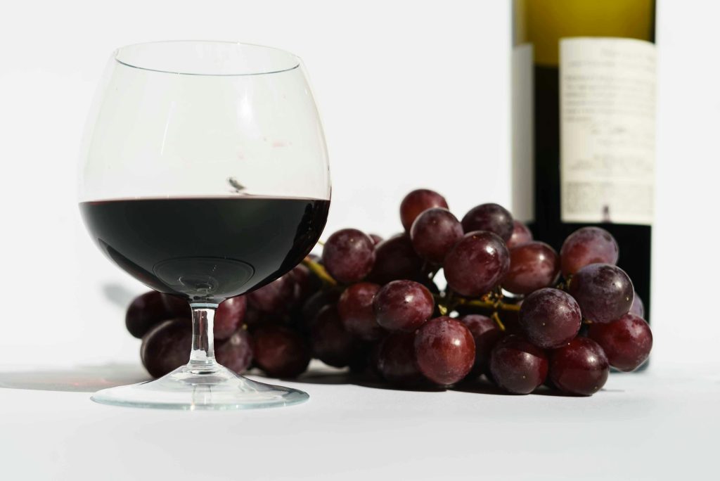 Glass of red fragrant wine placed near bunch of sweet grapes and bottle in white studio