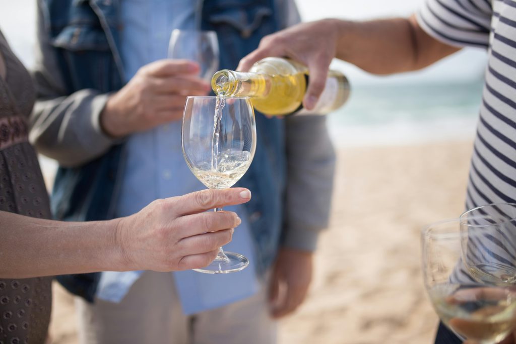 Wine and Steak Pairing: Best Bottles for Every Cut A person pours Pinot Grigio into a glass held by another on a sandy beach, with two others holding glasses in the background. The group stands close together, dressed casually, enjoying a relaxed wine comparison by the shore.