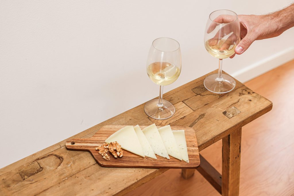 A wooden board with sliced cheese, walnuts, and white wine glasses on a rustic table.