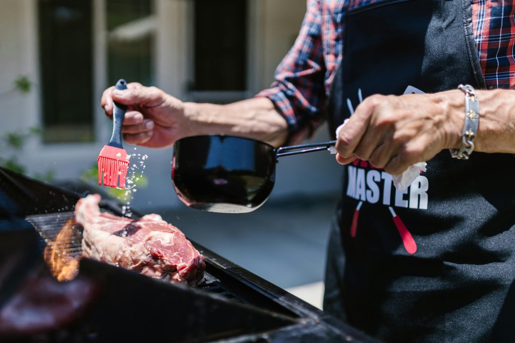 How to Pair Wine With BBQ: 5 Simple Rules A person prepares a juicy steak on an outdoor grill, using a silicon brush to apply sauce.