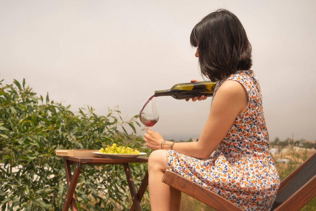A woman wearing a floral dress pours wine into a glass while sitting outdoors on a sunny day.