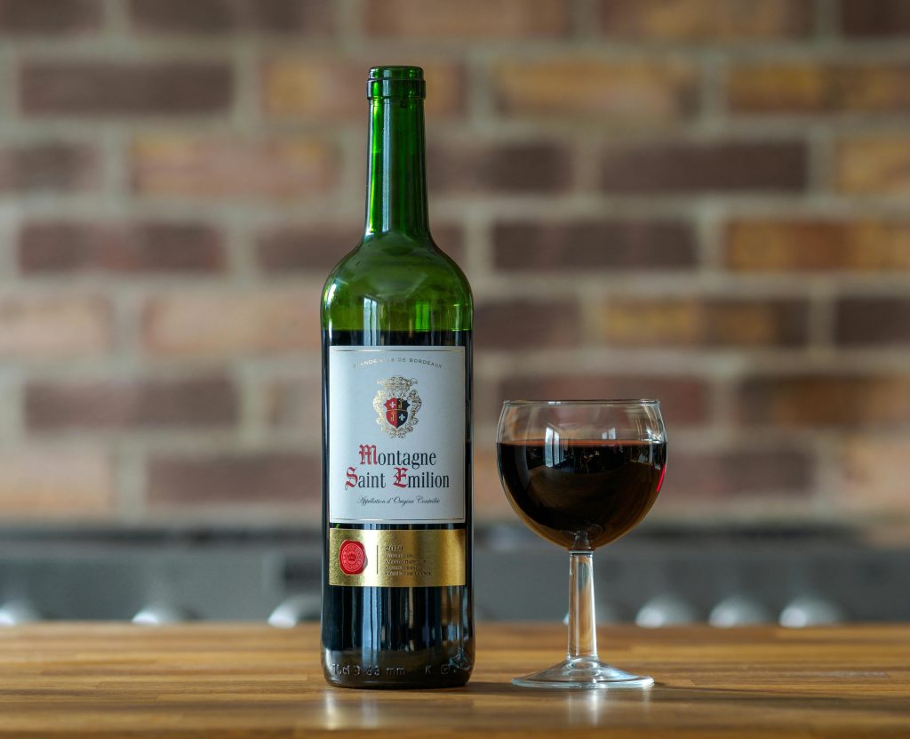 Best Bordeaux Wines Under $50 Close-up of a Montagne Saint Emilion wine bottle with a glass on a wooden table against brick wall.