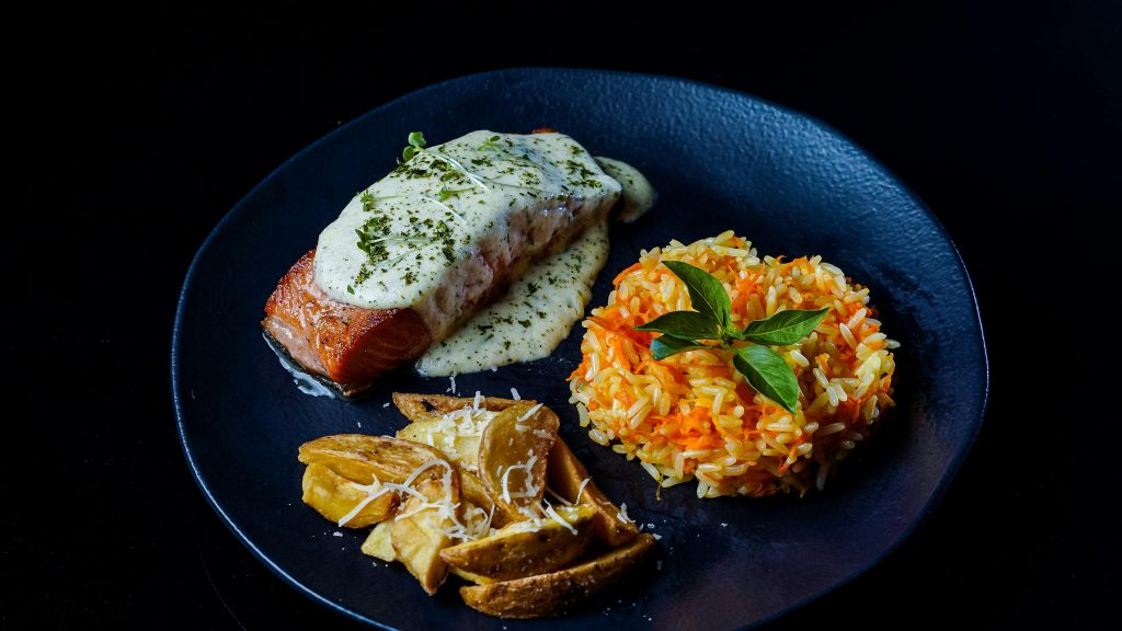 Wine and Salmon Pairing: The Complete Guide Delicious salmon served with creamy sauce, rice pilaf, and potato wedges on a black plate.
