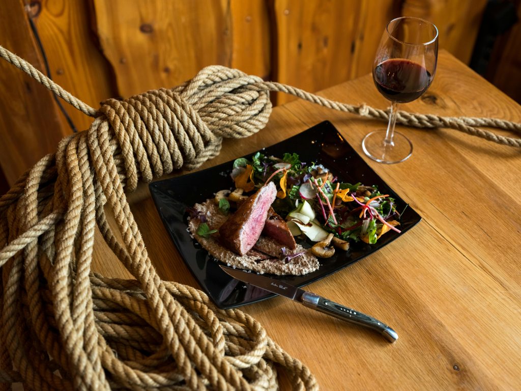 Wine and Steak Pairing: Best Bottles for Every Cut From above of ceramic plate with beef steak pieces topped on mushroom sauce and vegetable salad served with knife and glass of red wine placed on wooden table with rope tied in knot