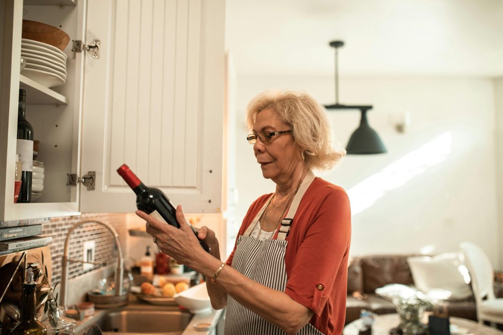 Elderly woman examining a wine bottle in a cozy kitchen setting.