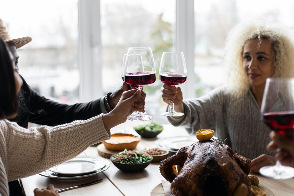 Happy diverse people clinking glasses with wine while sitting at table with roasted turkey and having Thanksgiving dinner