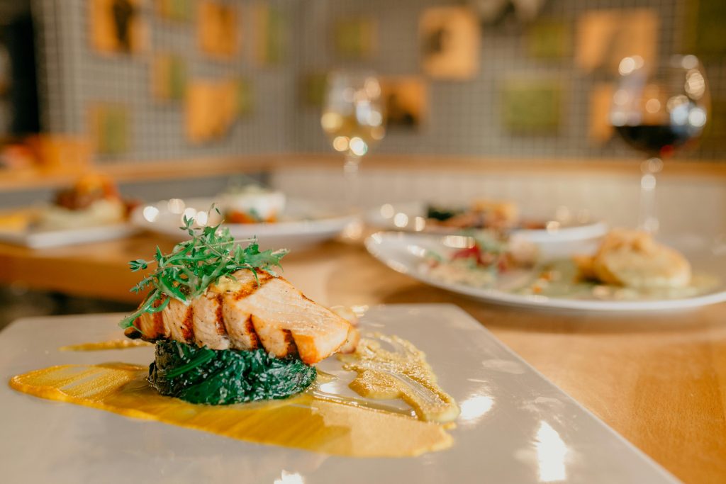 Wine and Salmon Pairing: The Complete Guide A plated dish featuring grilled salmon atop sautéed greens with a yellow sauce, garnished with microgreens. Blurred plates of food and glasses highlight an inviting wine and salmon pairing on a wooden table.