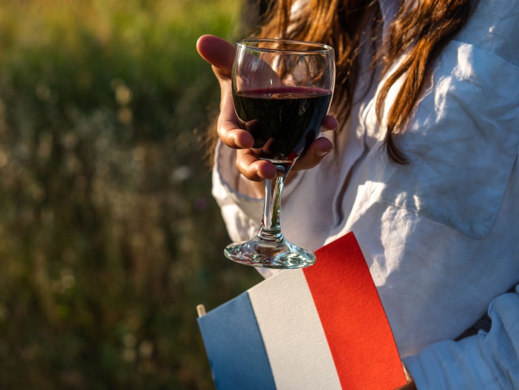 A person in a white shirt holds a French flag and a glass of red wine outdoors.