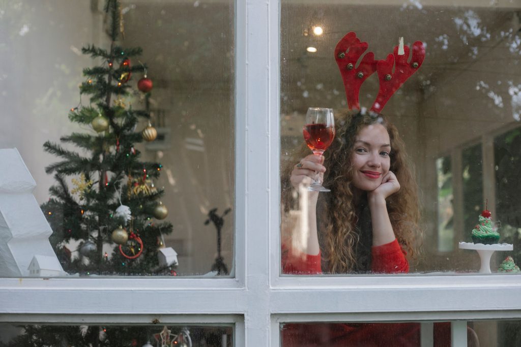 Best Wines for Holiday Hosting: Your 2026 Guide Through window of happy female in festive clothes sitting with glass of wine in room with Christmas tree and looking at camera
